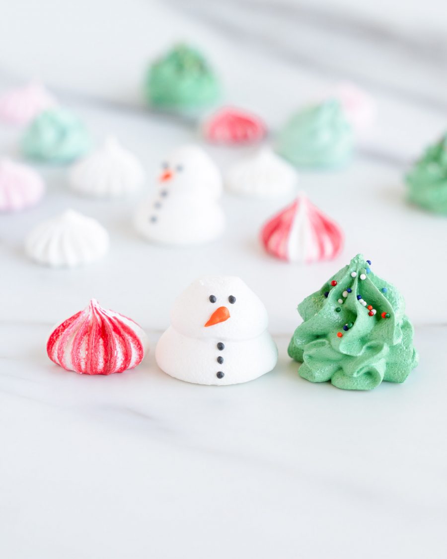 Holiday-Themed Meringues in Clear Round Container A clear round container filled with holiday-themed meringues, including snowman shapes, Christmas tree designs, and swirled red, green, and white meringue kisses.